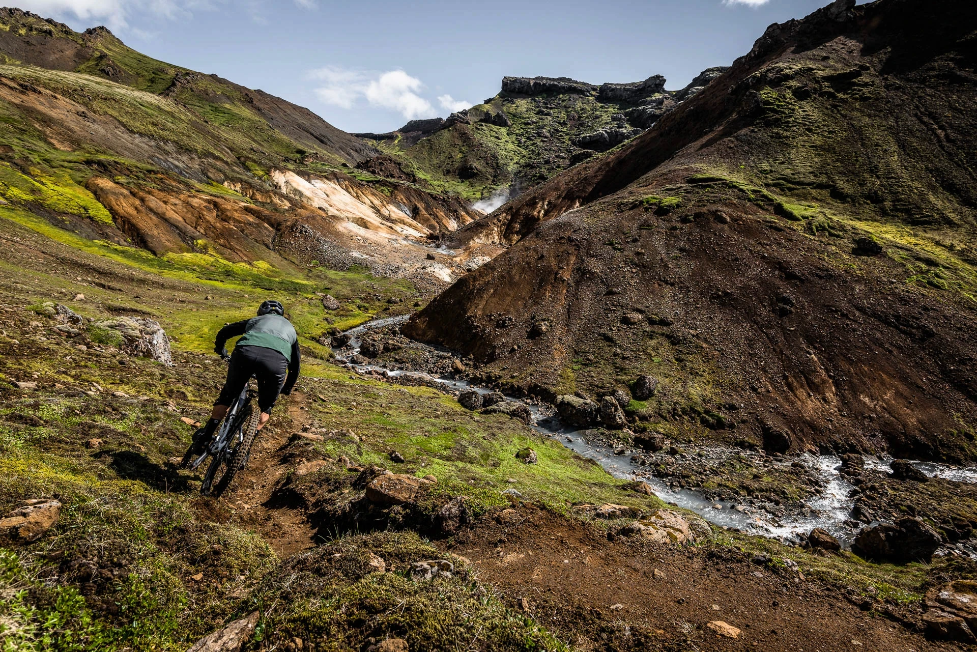 Mountain biker riding a trail beside a stream through a volcanic valley.