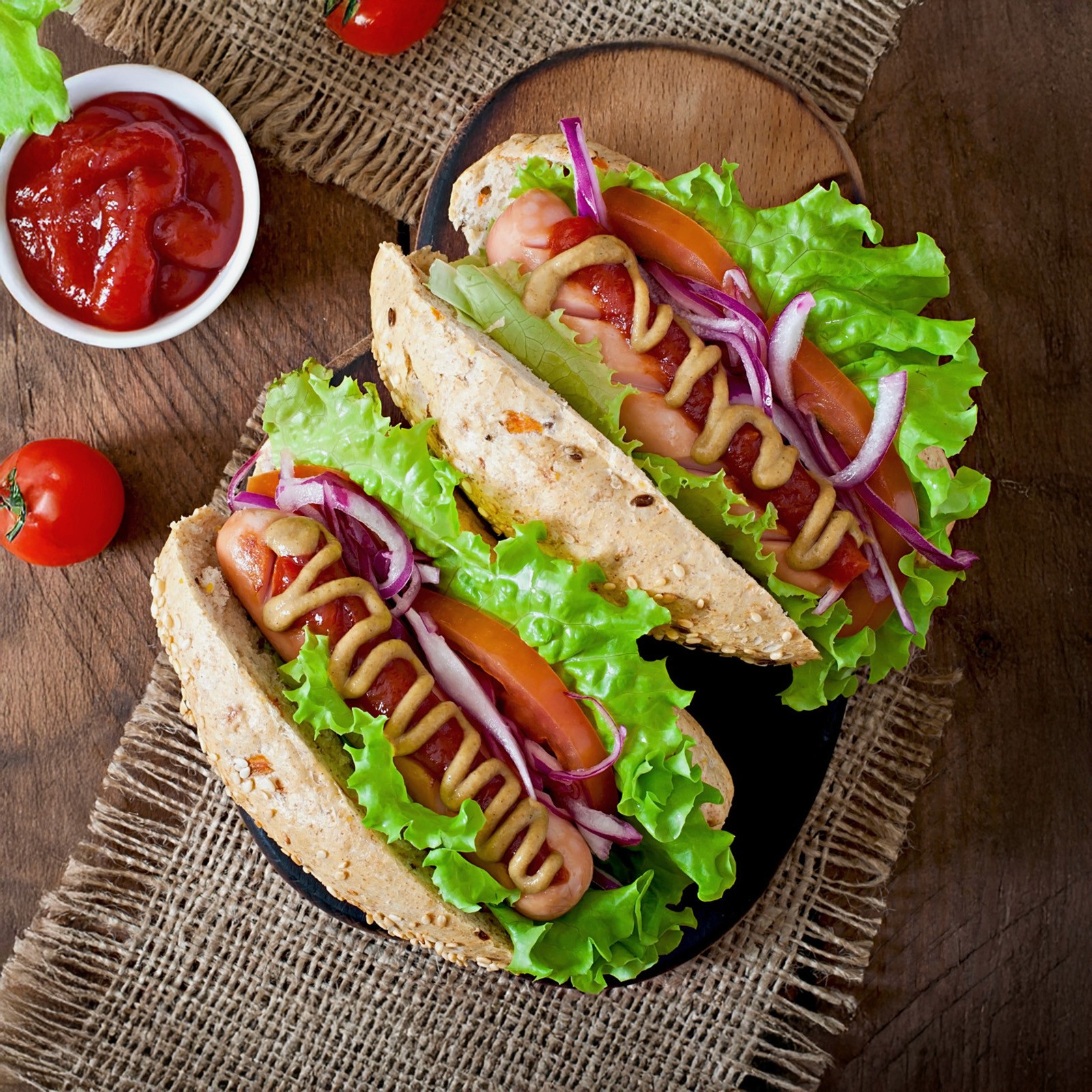 Top-down view of two loaded hot dogs in seeded buns with lettuce, tomato, red onion, mustard, and ketchup, served on a wooden board over a rustic burlap cloth.