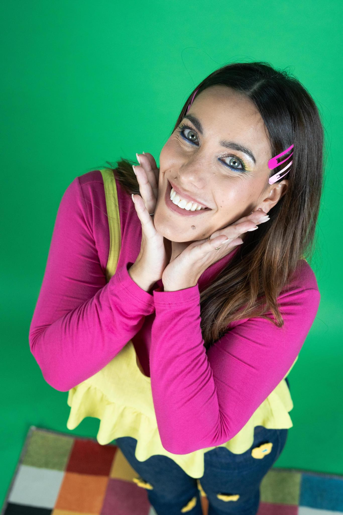A woman with blonde bob hair in a grey coat and beige turtleneck stands against a neutral backdrop. She has a serious expression, conveying elegance.