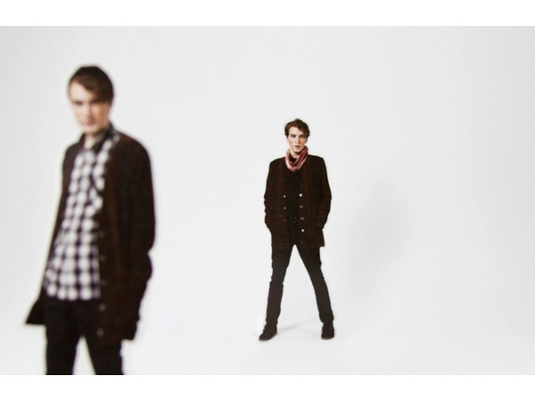 The image shows two young men against a plain white background. The man on the left is out of focus, wearing a black and white plaid shirt under a dark brown jacket and dark pants. His hands are in his pockets. The man on the right is standing more centrally, wearing a dark brown cardigan over a dark shirt, a red scarf around his neck, dark pants, and dark shoes. His hands are in his pockets. Both men have light-colored hair.