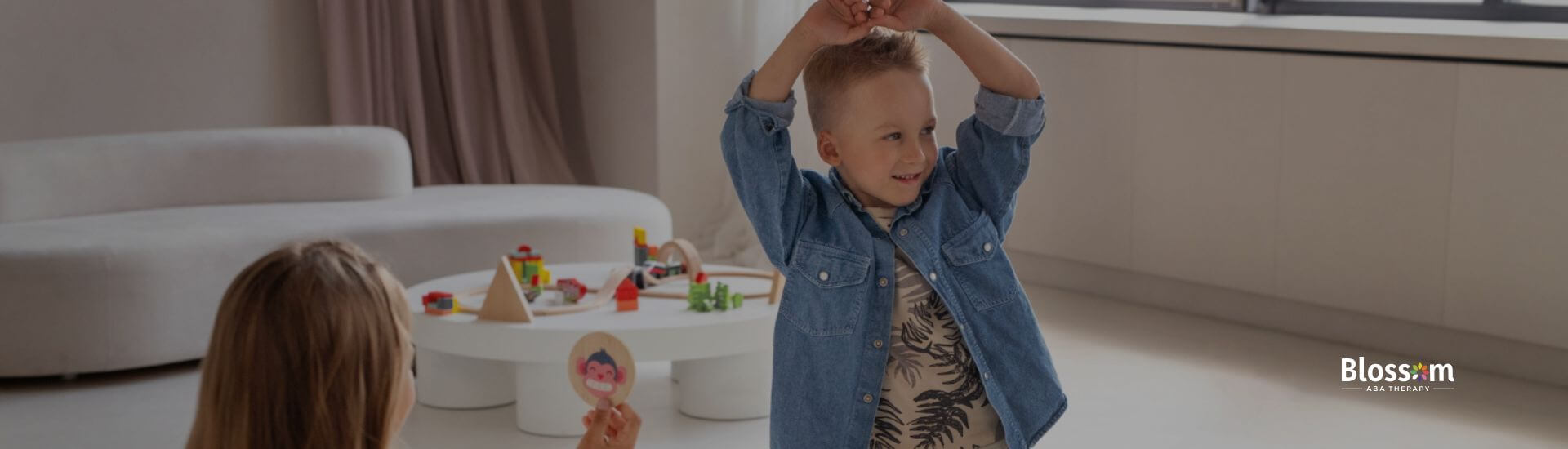 Child with autism raising arms during an ABA therapy activity while the therapist holds a picture cue.