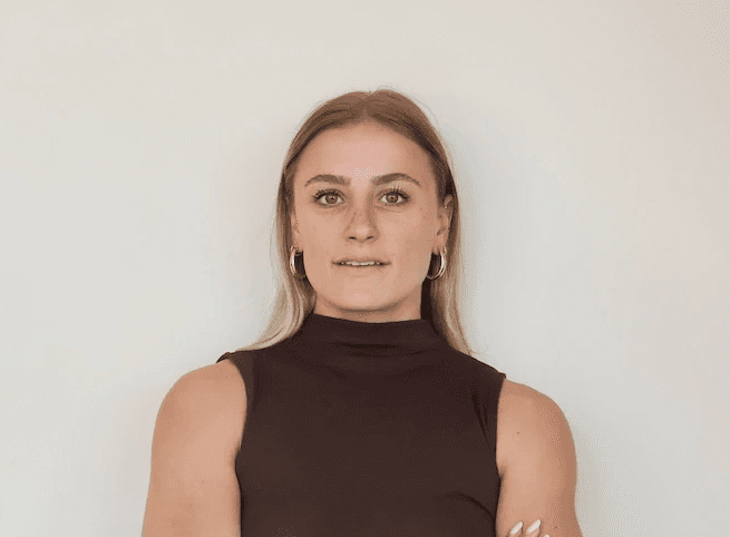 A young man with light brown hair is wearing a black suit jacket and white shirt, standing in a softly lit office setting, conveying professionalism.
