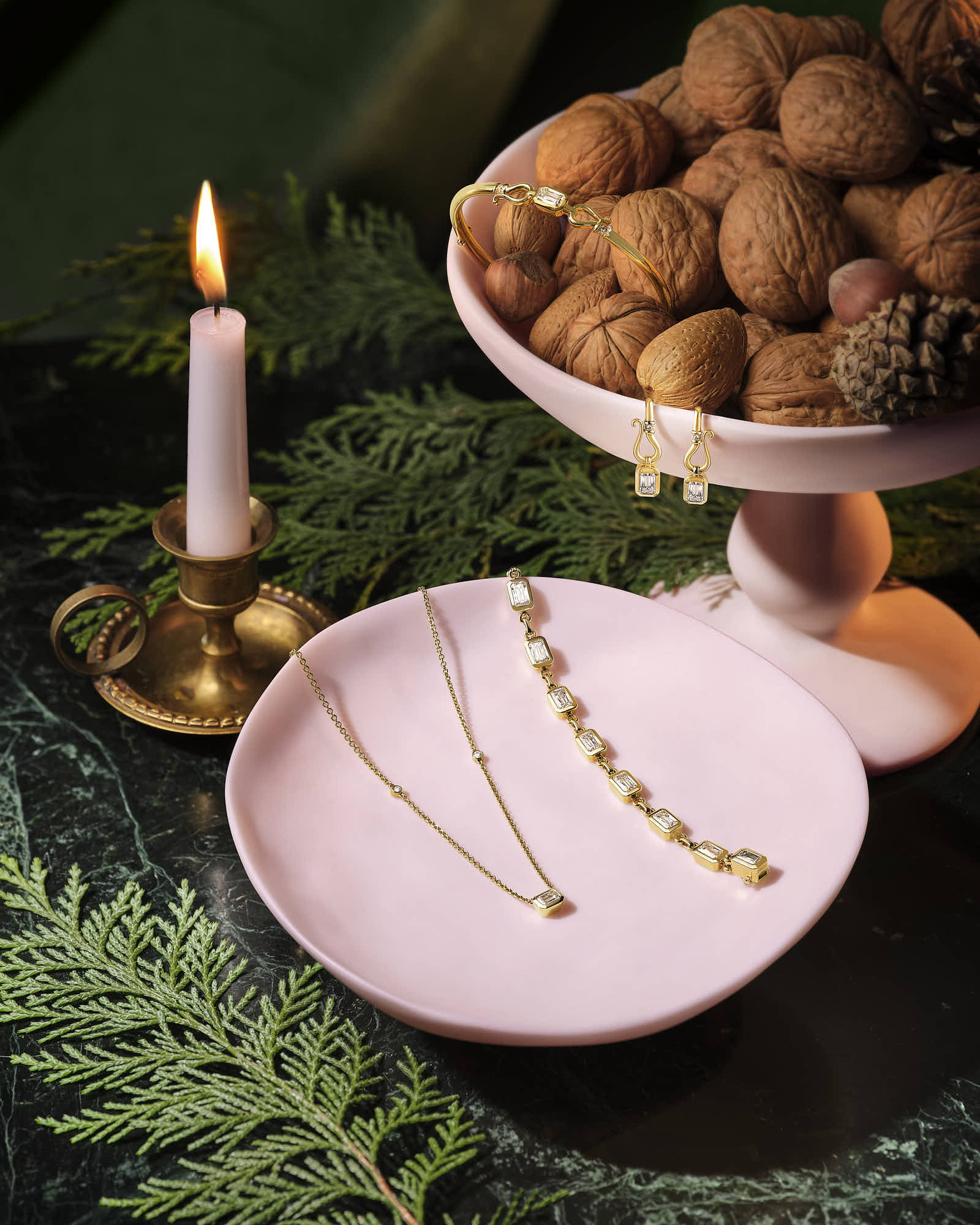 A decorative setup featuring jewelry on a plate, a bowl of fruits, and a lit candle against a dark background.
