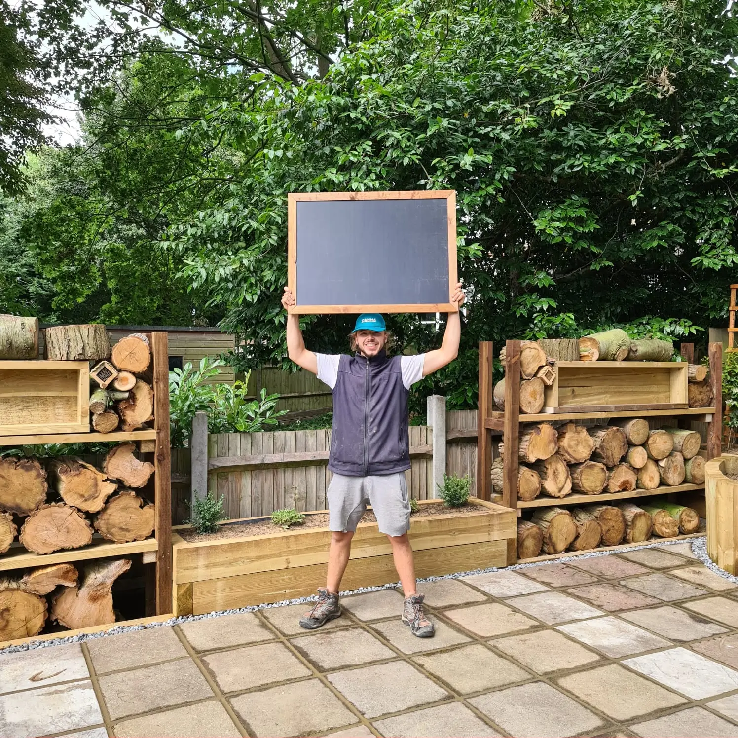 A person stands in a yard, holding a sign overhead, surrounded by stacked wooden logs and greenery.