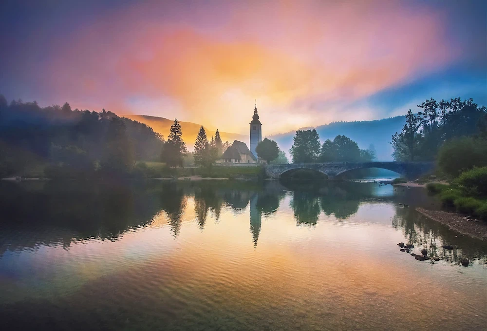 A church reflecting in Lake Bohinj in Slovenia during pink foggy sunrise.