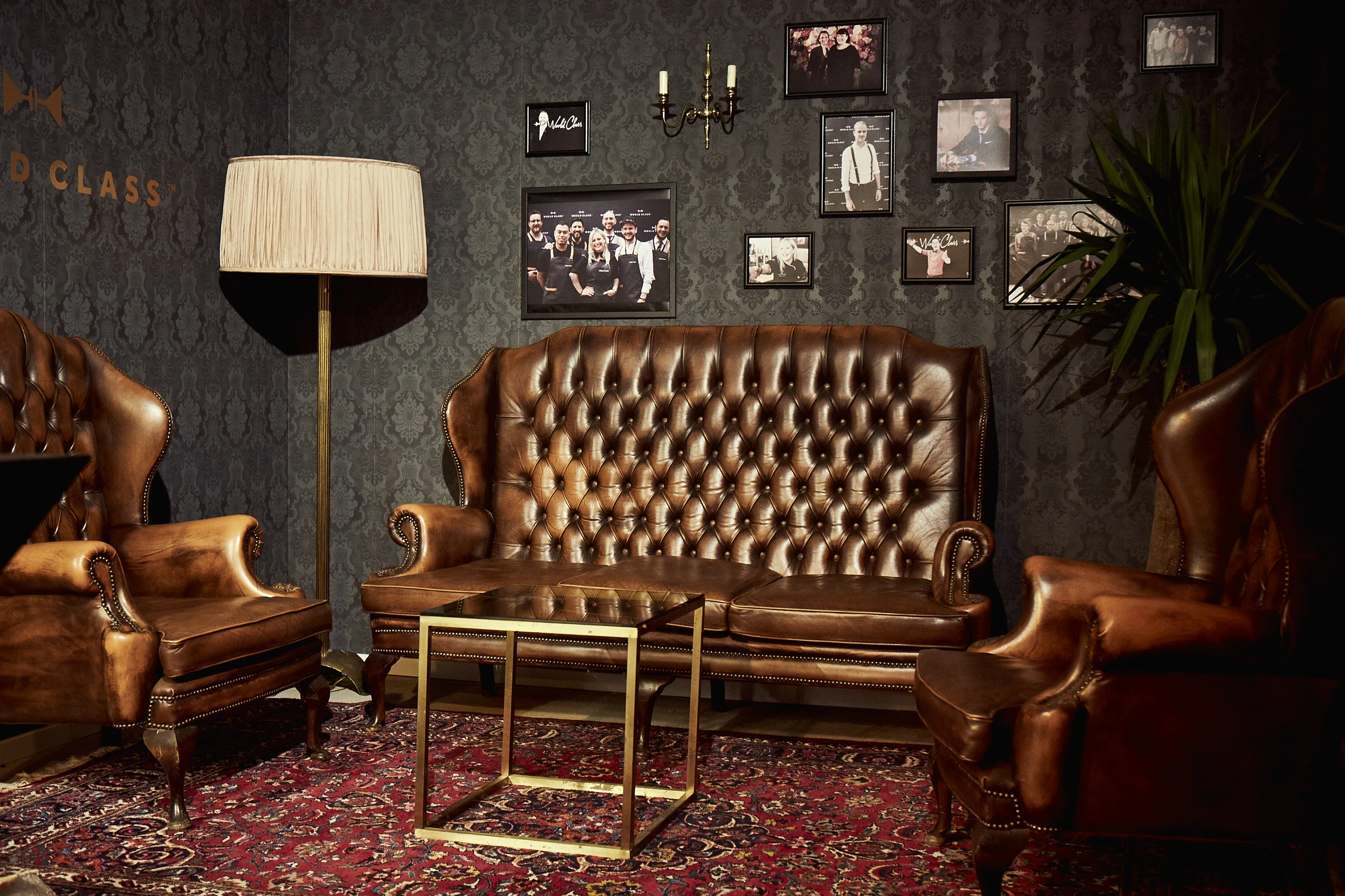 Gentleman-style lounge at DIAGEO BCB17 with brown Chesterfield sofa and wing chairs, brass cube table, framed photos on dark patterned wallpaper and a Persian rug.