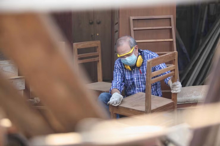 A man sanding a wooden chair in a workshop, wearing gloves, a cap, and safety earmuffs.