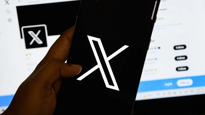 A hand holding a smartphone displaying the bold X (formerly Twitter) logo, with a blurred background showcasing the X interface.