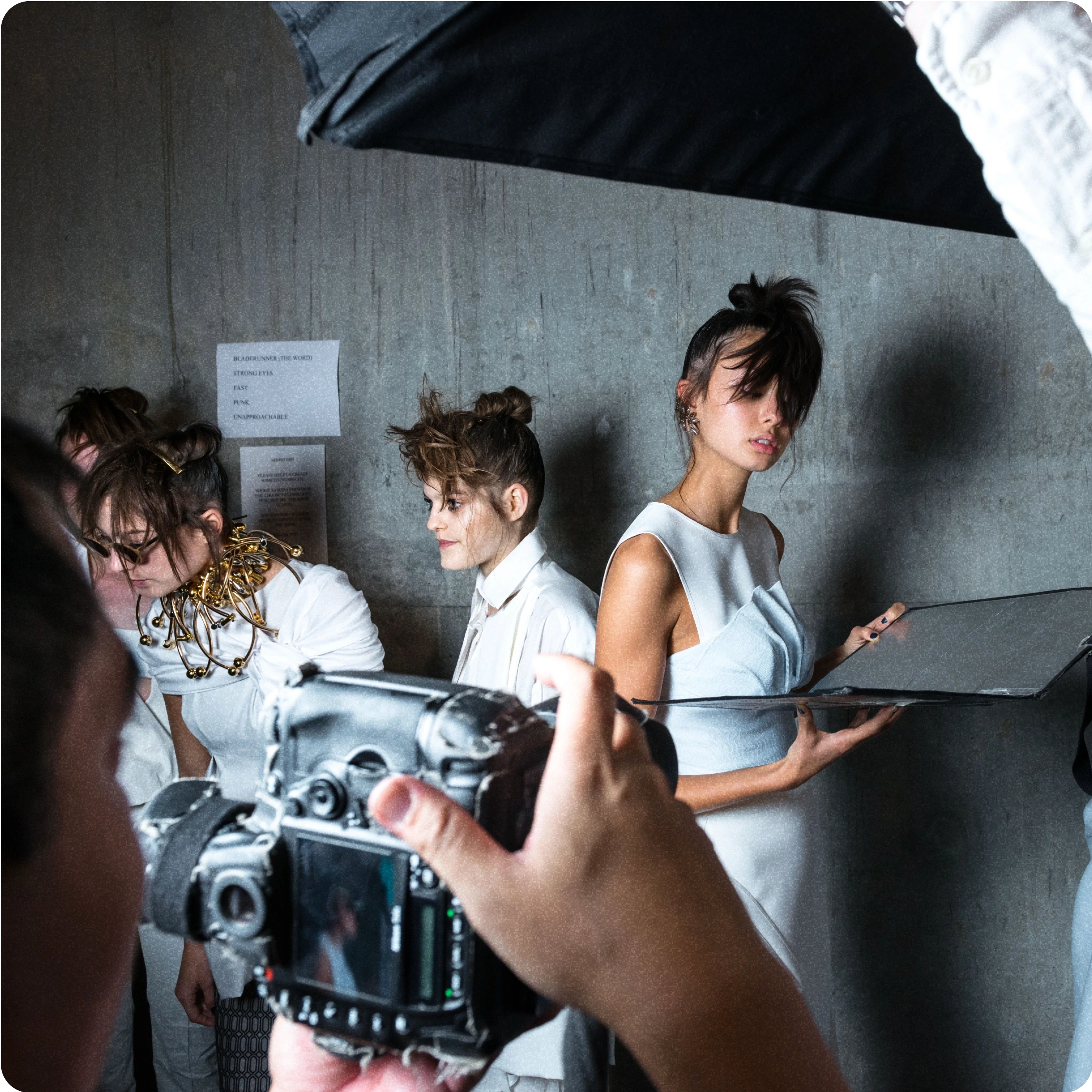 A behind-the-scenes shot of a fashion photo shoot. Three models are positioned against a grey concrete wall, wearing avant-garde white outfits with creative hairstyles. A photographer's hands and camera are visible in the foreground, capturing the scene. One model is holding a reflector, aiding the photo shoot setup. The setting is vibrant and dynamic, showcasing the fashion photography process.