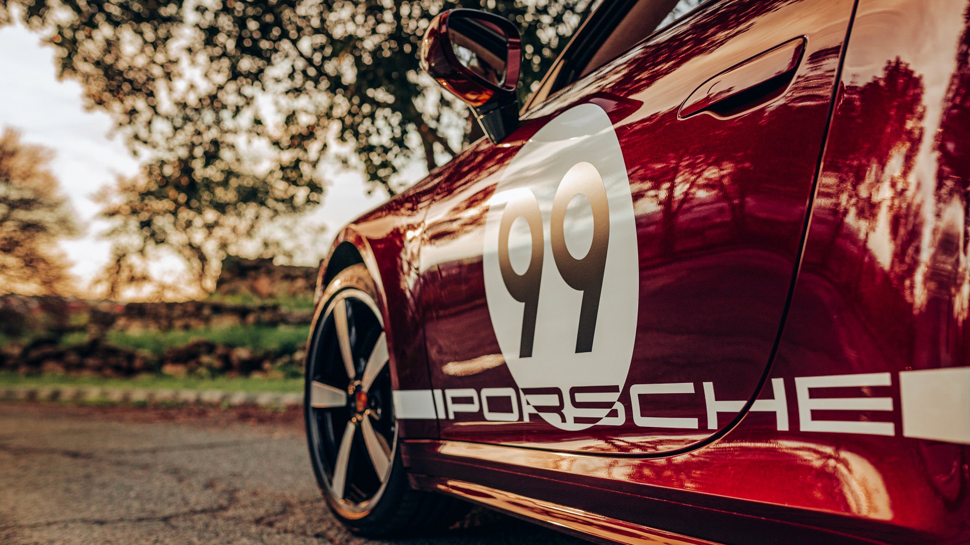 Side detail of a red Porsche 911 Targa 4S Heritage Design Edition, low angle on the white "99" roundel and gold "PORSCHE" script along the door, trees reflected in the deep cherry paint.