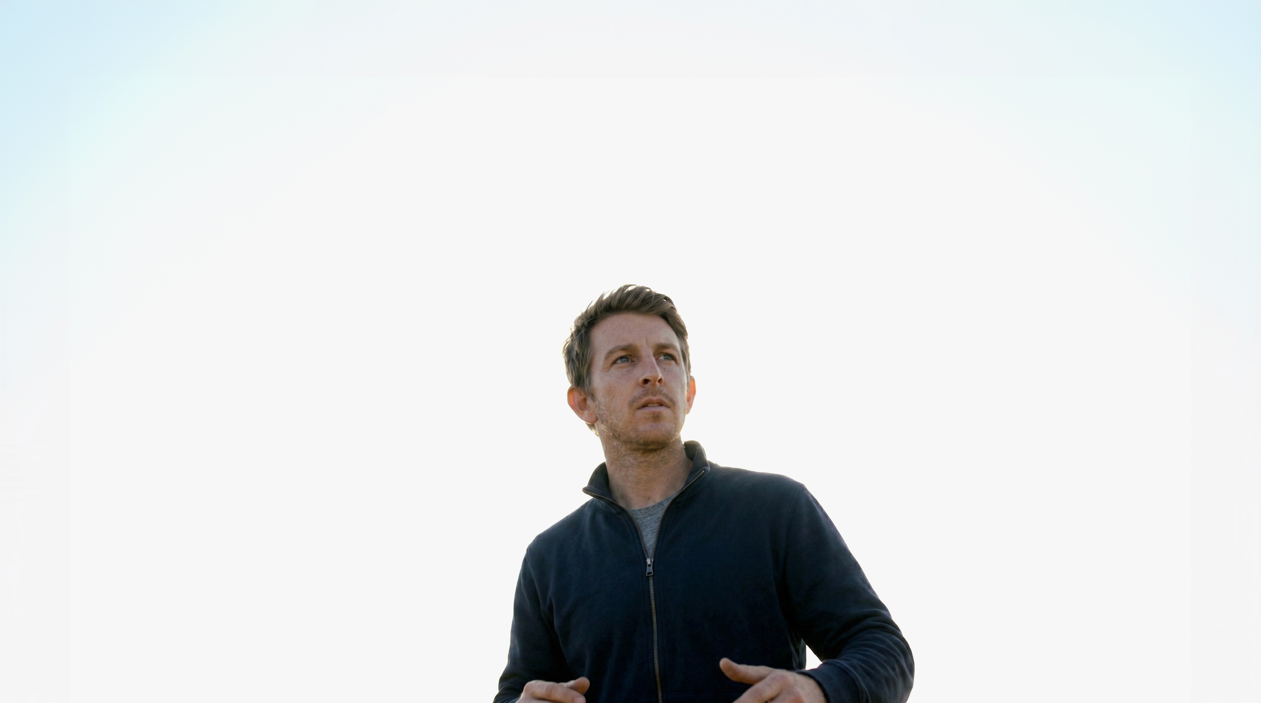A man in a dark jacket stands outdoors against a bright, clear sky, looking upwards with a thoughtful expression.