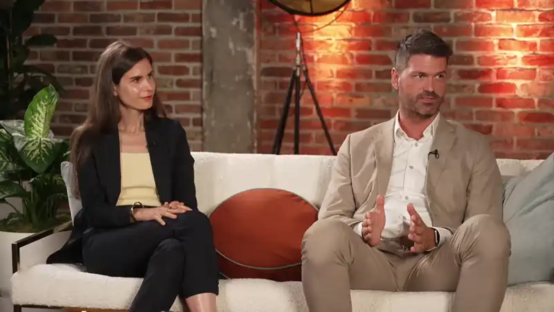 Two panel speakers seated on a studio sofa during a live discussion, framed against a brick backdrop