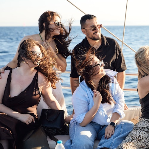 Four people enjoying a sunny day on a boat, with wind blowing their hair, against a backdrop of a calm sea.