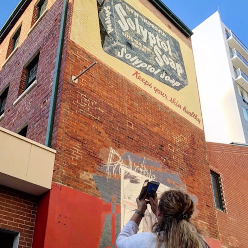 A person takes a photo of a mural on a brick building that advertises Solvol soap. A modern building is visible in the background.