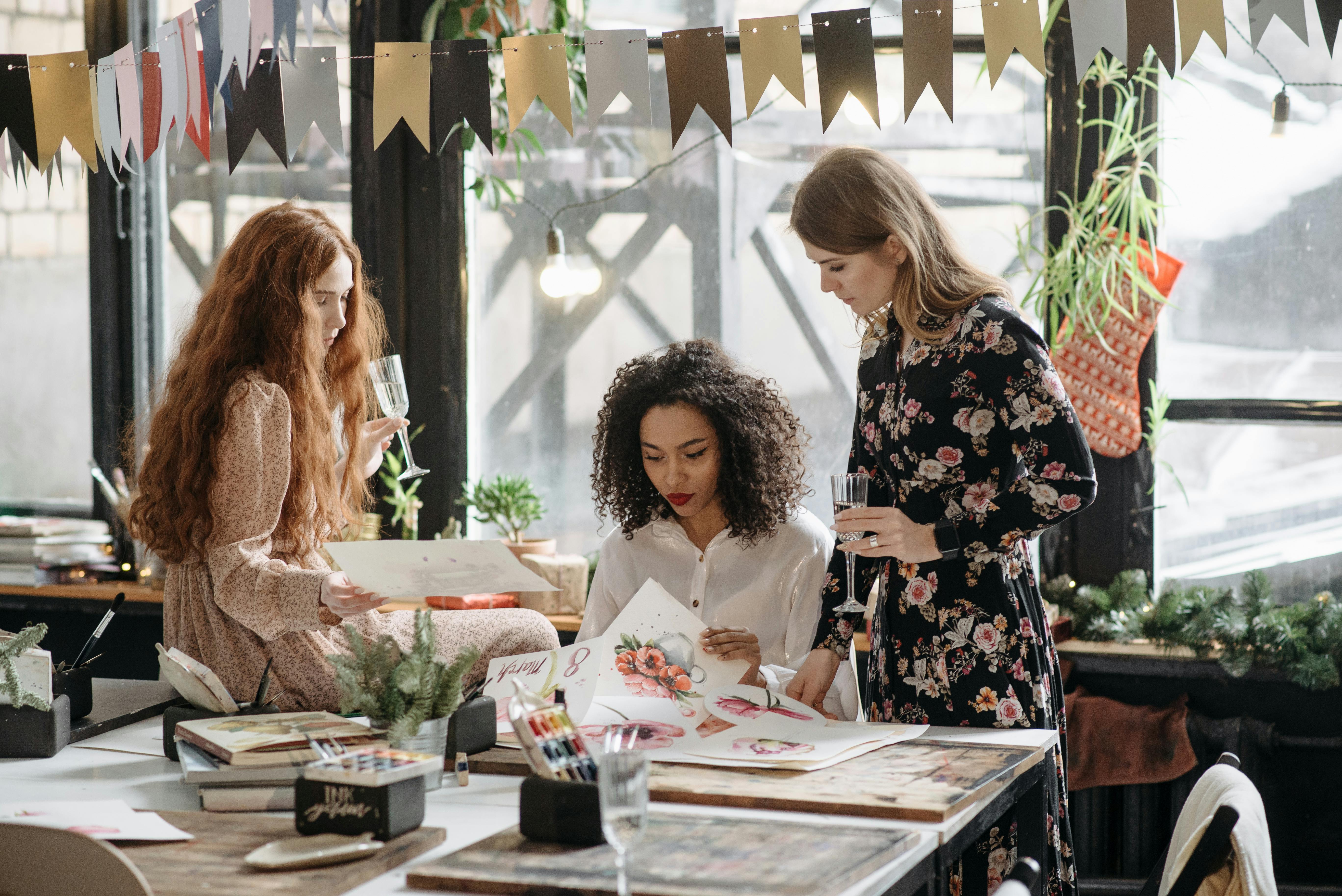Drei junge Frauen in einem kreativen Workshop betrachten gemeinsam Aquarellbilder an einem Tisch, trinken aus Gläsern, lachen und gestalten, Symbol für Freundschaft, Brautparty und kreative Mädelsaktivitäten.