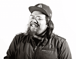 A black and white portrait of a man with long dark hair and a beard, wearing a dark jacket and a baseball cap with a logo. He is smiling and wearing glasses. The background is plain white.