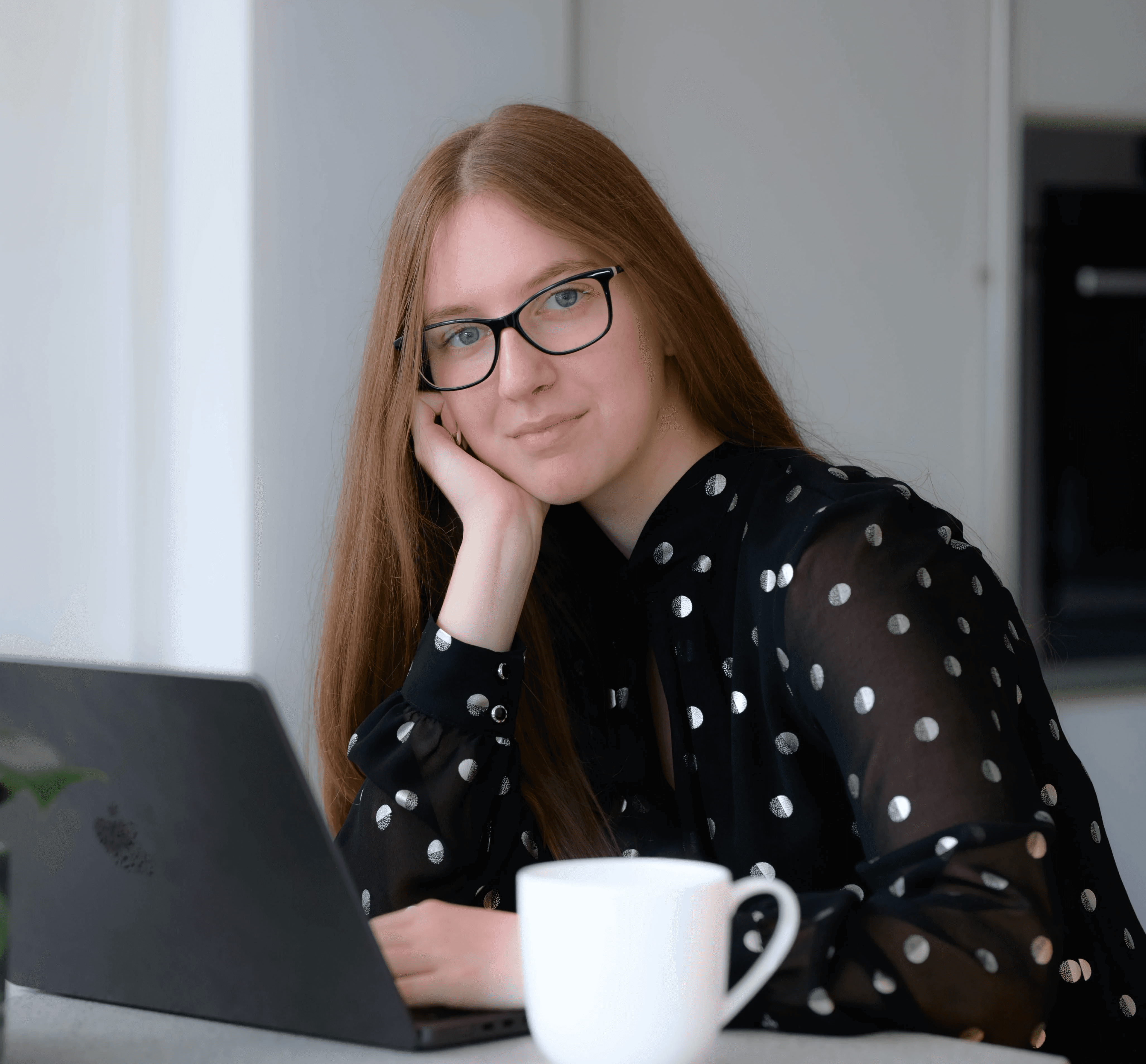 A young person with curly hair smiles at the camera while sitting in front of a laptop against a dark background.