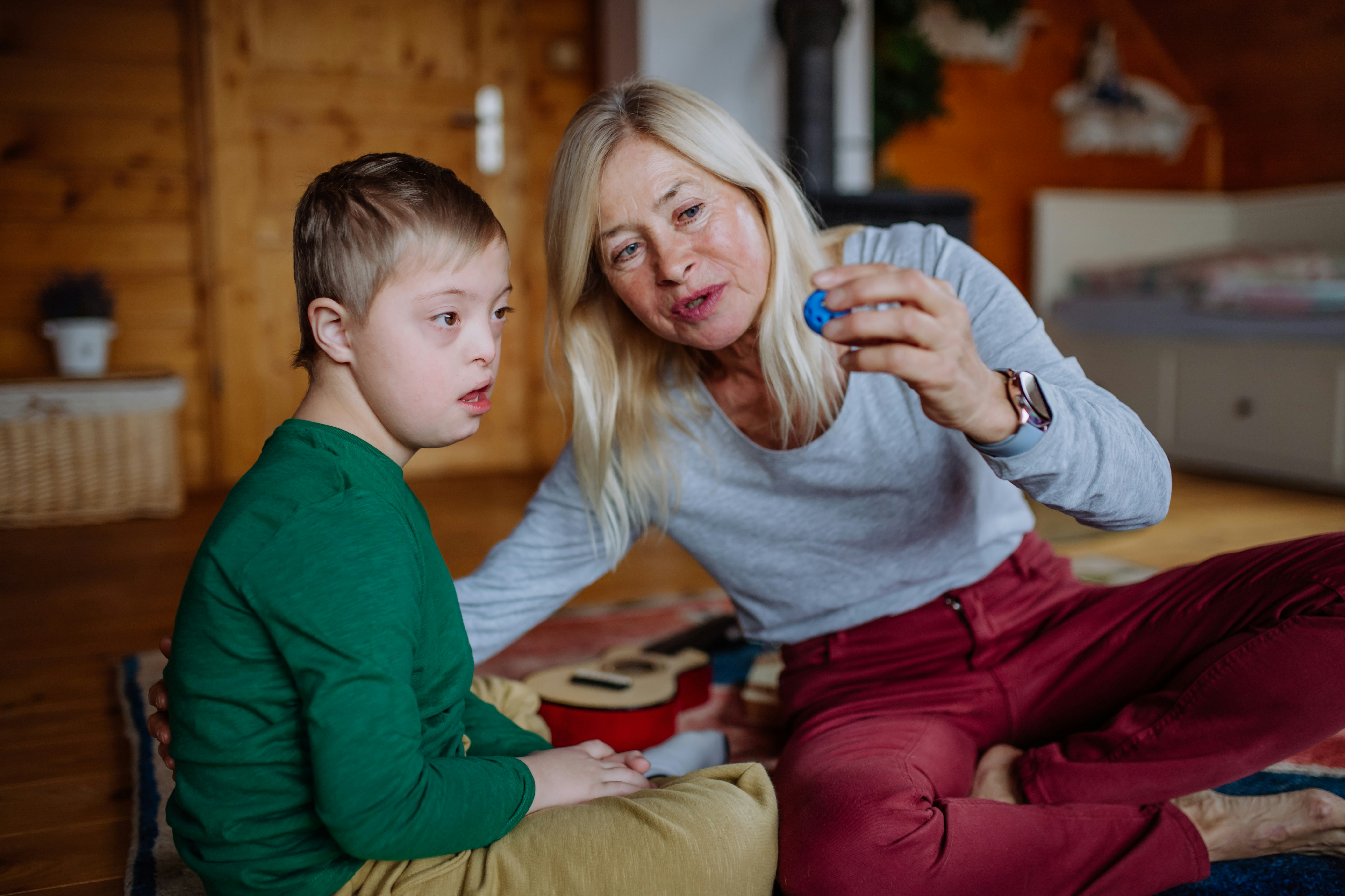 A woman and a boy sitting on the floor of a room with wood paneling, the woman holding a small blue object.