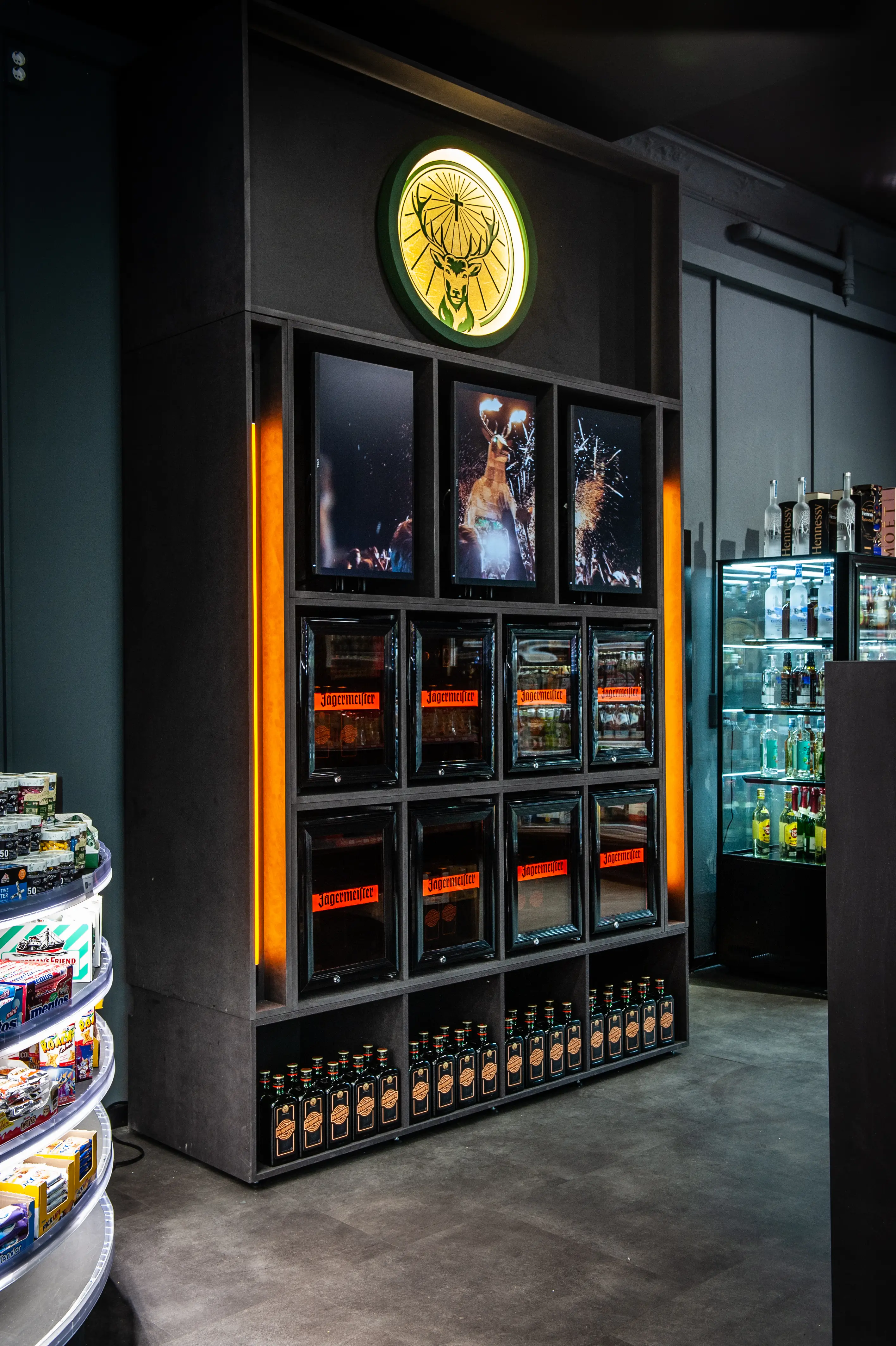 Black fridge wall unit at JÄGERMEISTER x SVENNI'S with a glowing green stag logo, screens, orange light edges and a row of Jägermeister bottles at the base.