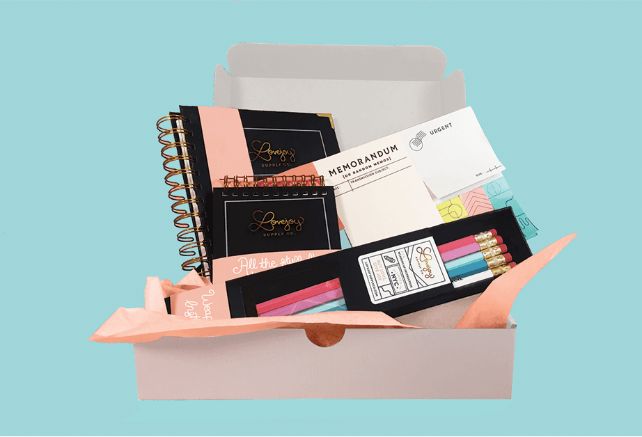 A white box contains various stationery items: a black spiral-bound notebook with “Lovejoy Supply Co.” branding, a white memo pad labeled “Memorandum for Randon Mendoza,” a black box labeled “All the studio,” a black box labeled “All the studio,” and a set of colored pencils in a black box labeled “Lovejoy Studio.” The box is set against a light blue background.