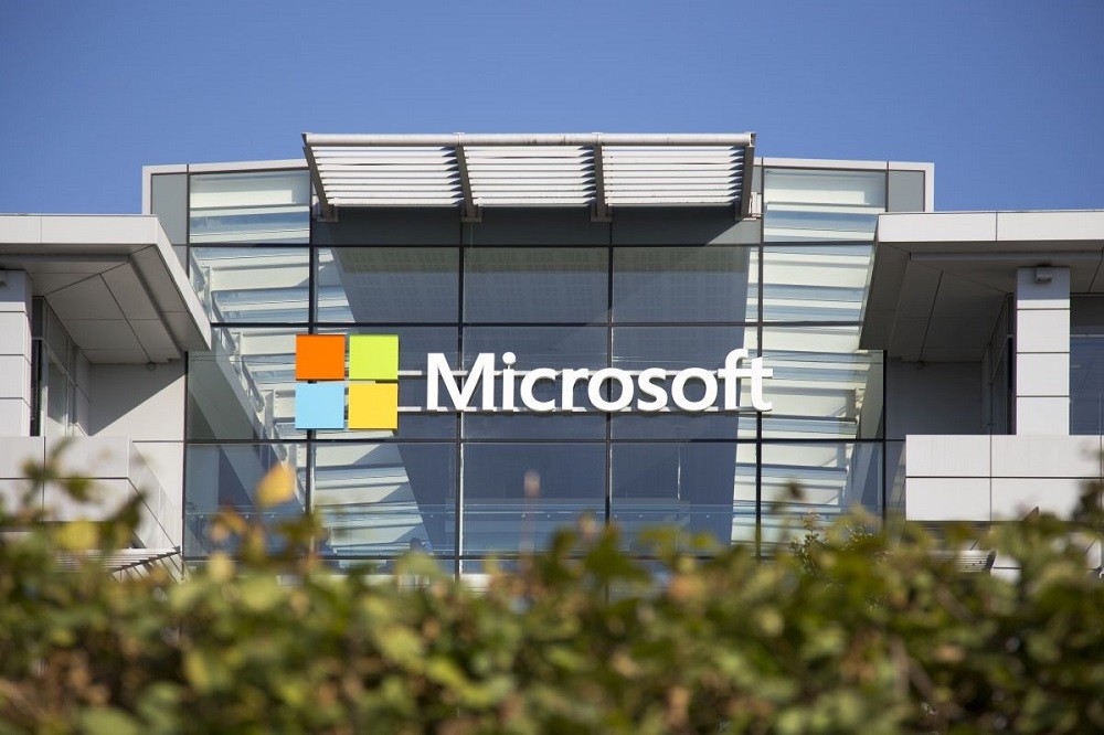 Microsoft building with a large, colorful Microsoft logo displayed on the glass facade, featuring modern architecture and greenery in the foreground.