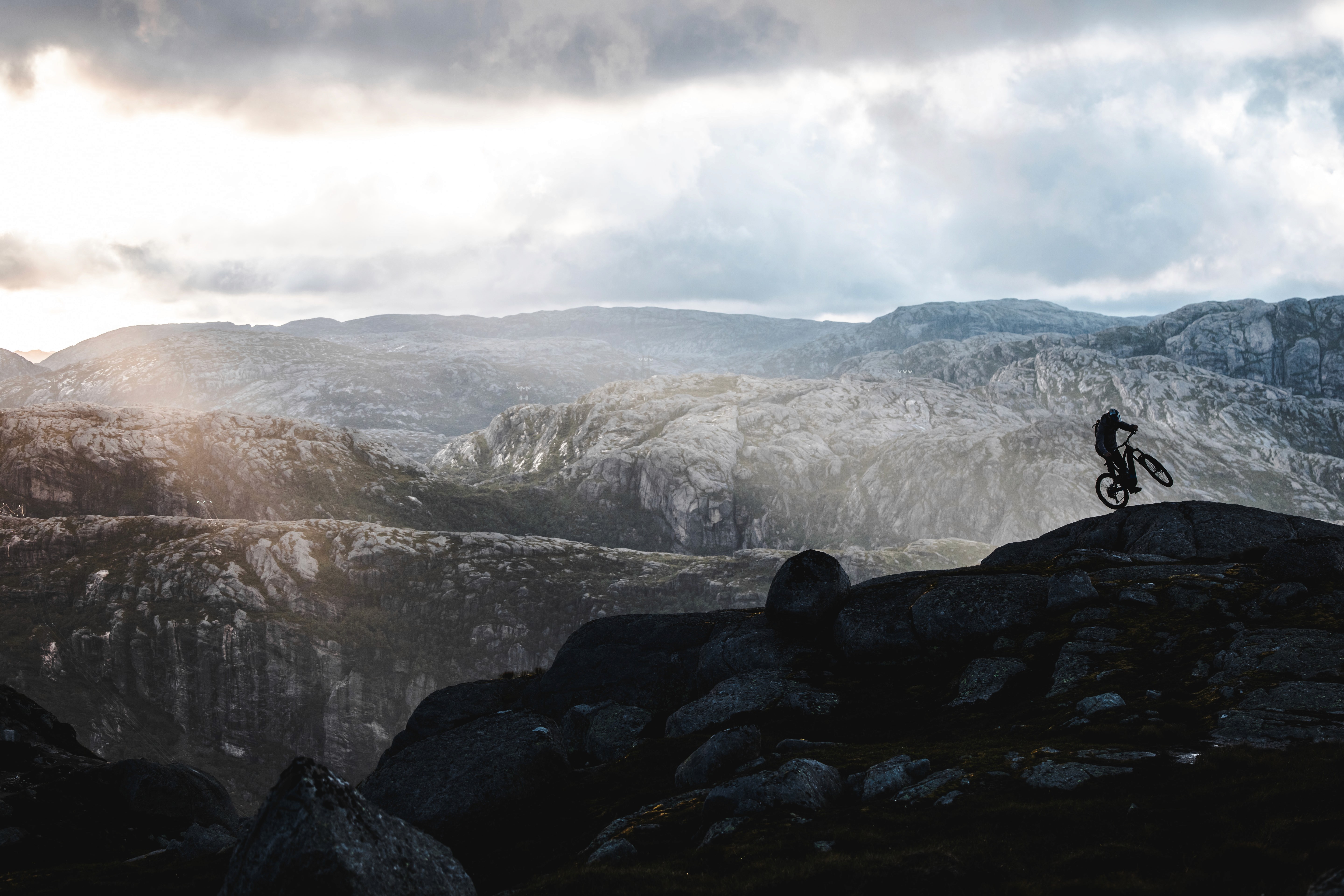 Silhouette of a mountain biker on a rocky ridge overlooking a vast, rugged Norwegian mountain landscape under a dramatic cloudy sky at dusk.