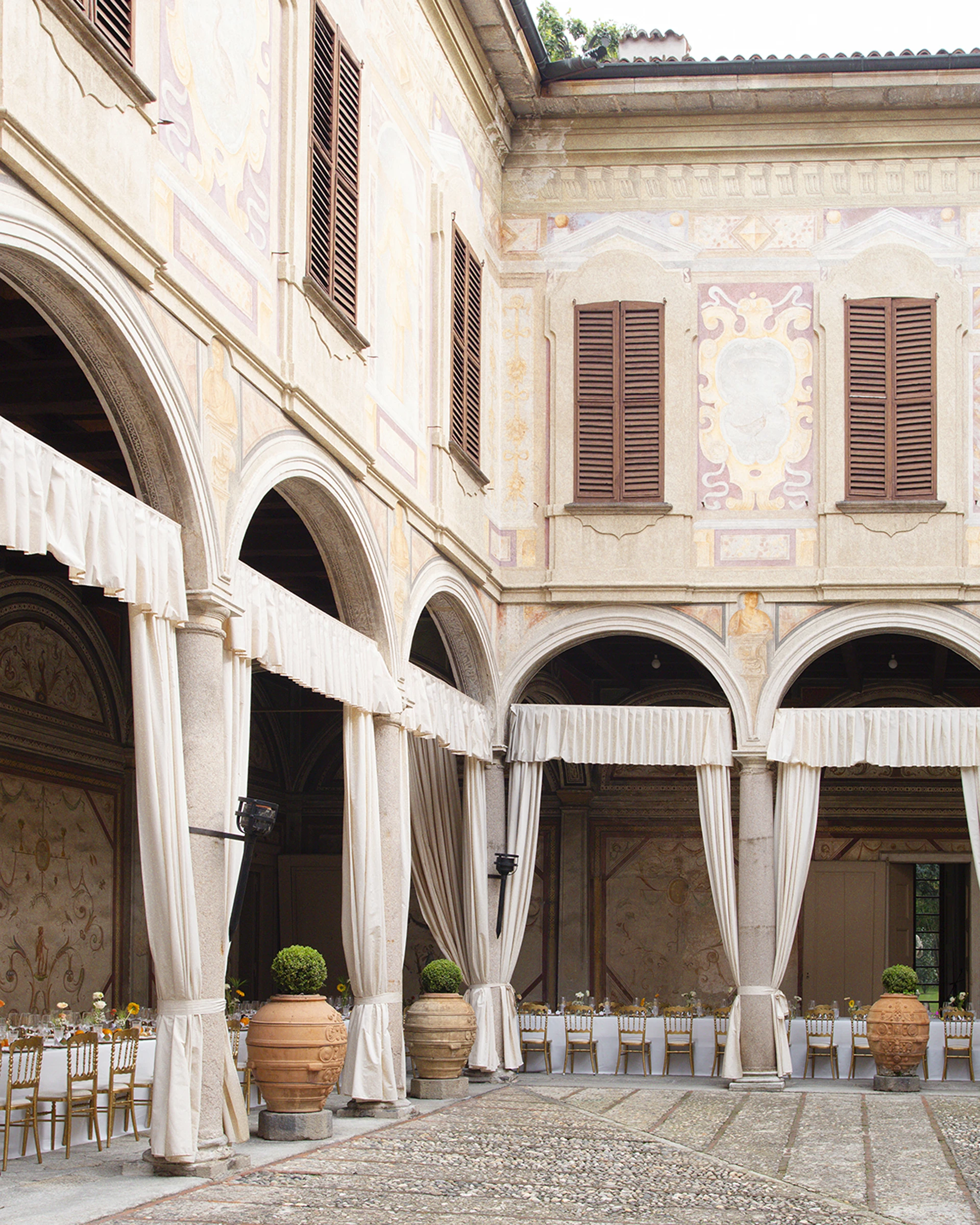 Cortile porticato con arcate, tende chiare e vasi in terracotta con piante sagomate