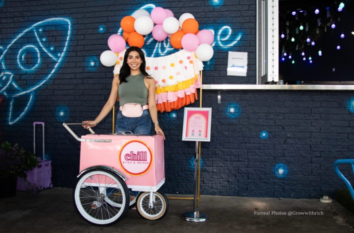 A polaroid photo of a woman standing behind a pink cart with a balloon archway behind her against a dark blue graffitied building