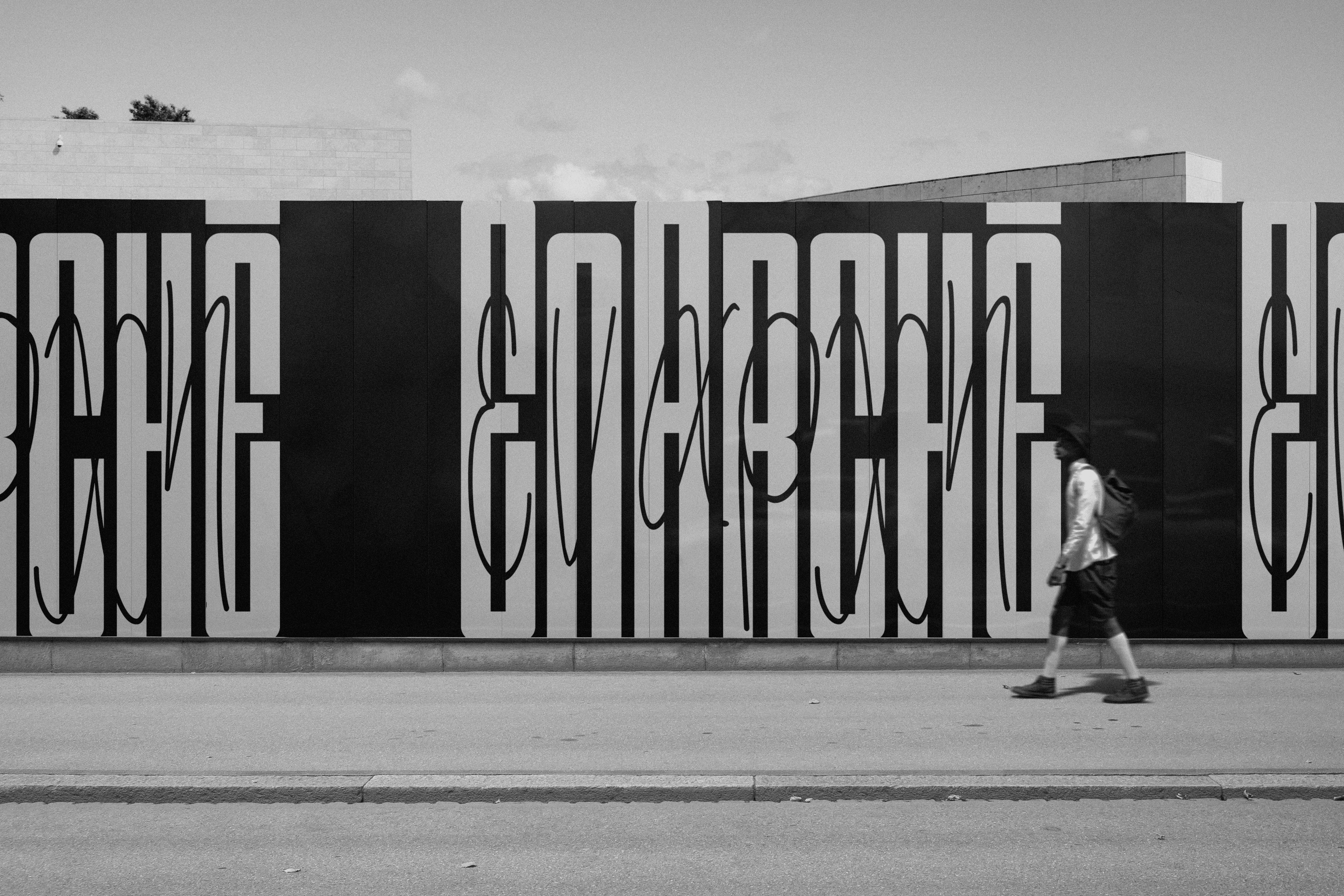 Tall fence branded with EnArchē's logo, partially occluded by a young man walking by.