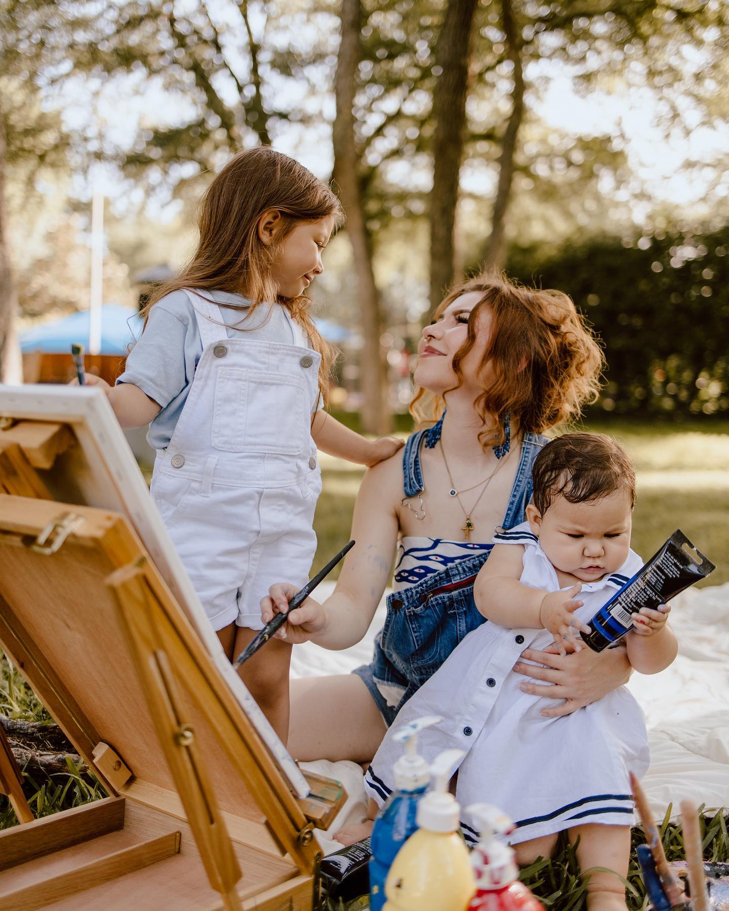 A young mother with red hair, wearing denim overalls, sits on a blanket and lovingly holds her smiling baby dressed in a sailor-style outfit. The lush greenery of a park with soft sunlight filtering through creates a warm, joyful scene.