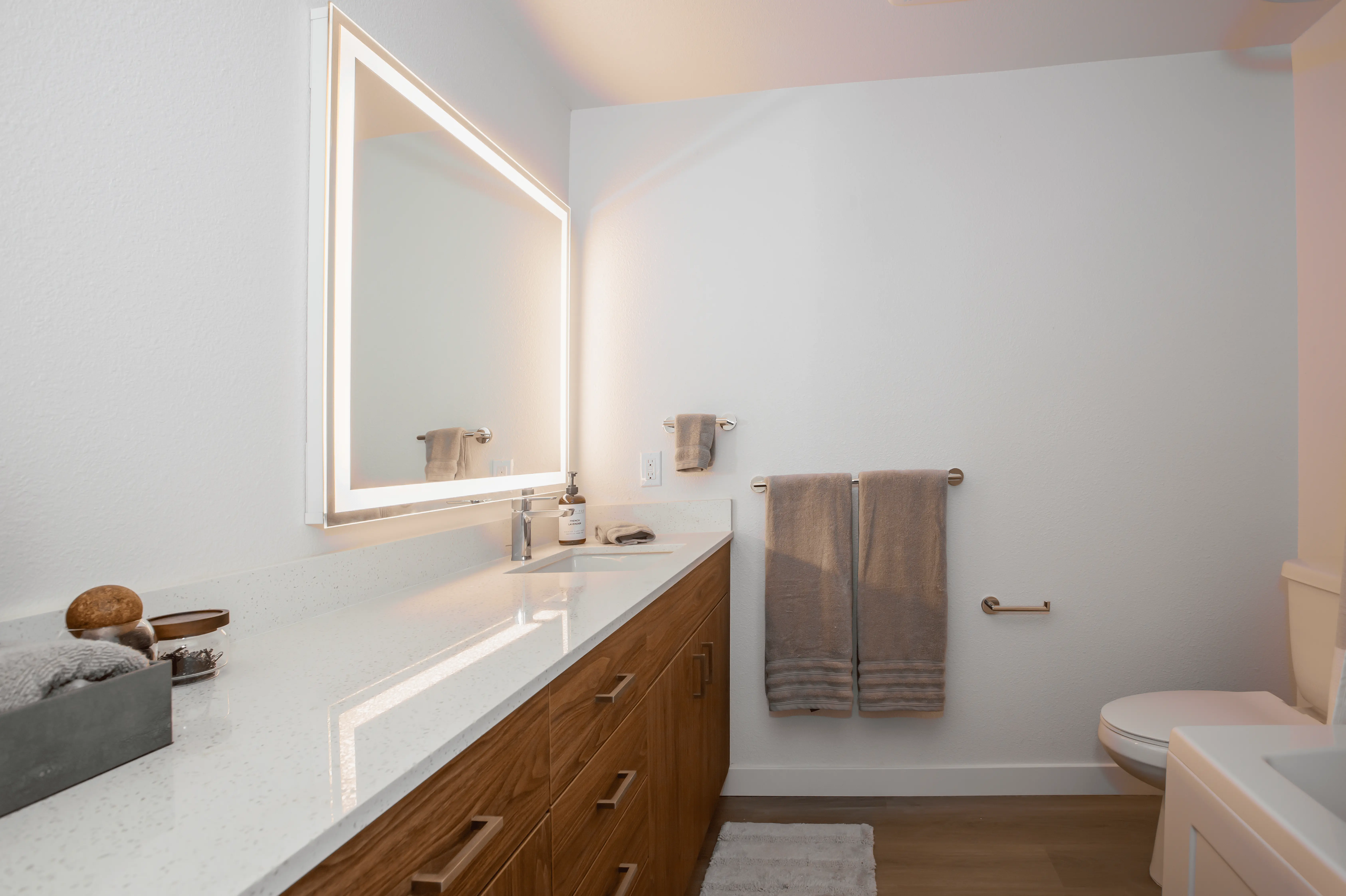 A modern, sun-drenched bathroom features a large vanity with a quartz countertop, wooden cabinets, and an illuminated mirror. Toiletries are neatly organized, and gray hand towels hang on the wall. The room also includes a toilet, toilet paper holder, and a soft rug on the floor—perfect for modern living spaces in Midtown Sacramento.