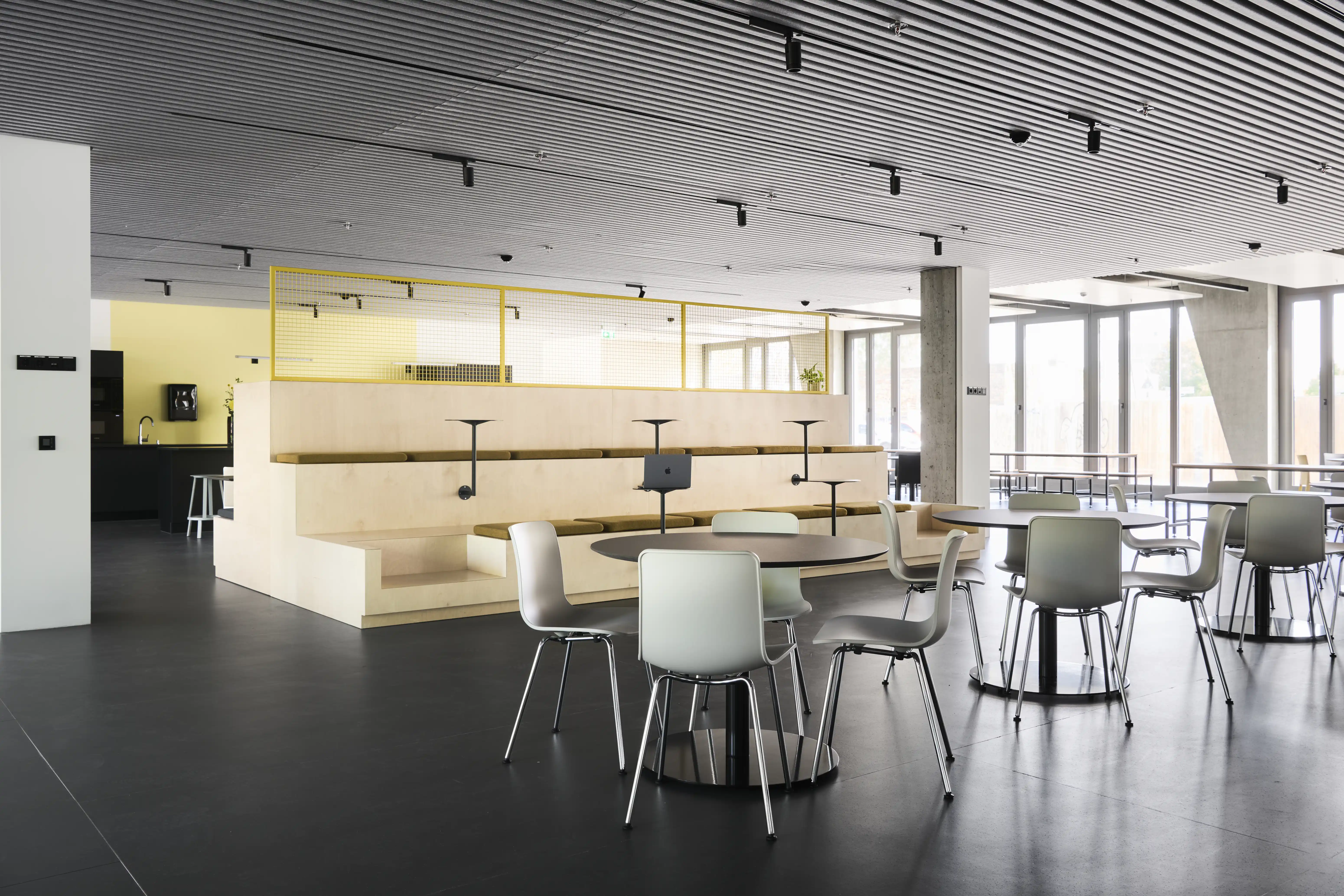 Cafeteria at ENPAL with round black tables and pale grey chairs in front of a birch seating tribune with yellow mesh railing and a ribbed metal ceiling.
