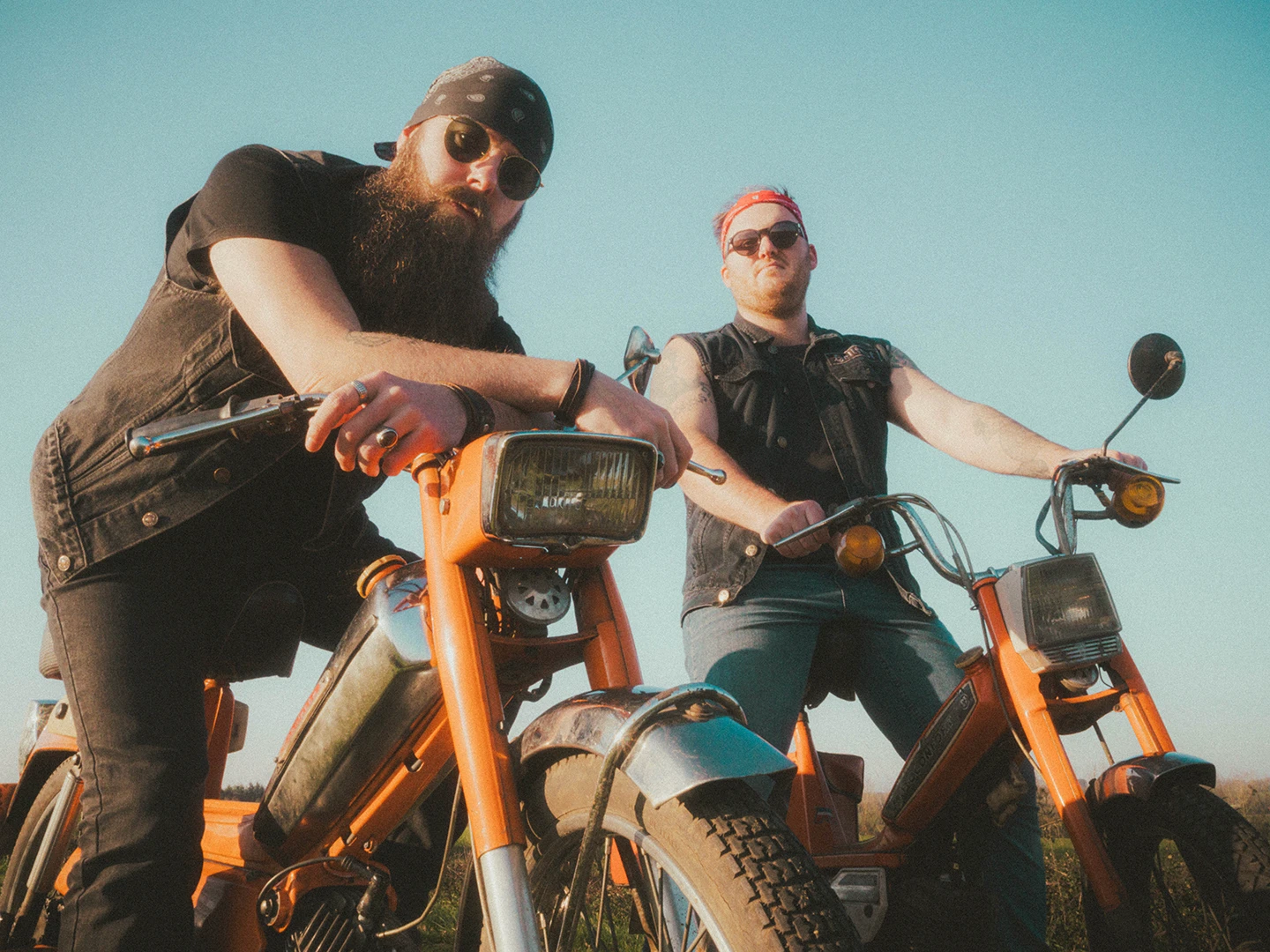 Deux hommes en contre-plongée en tenue de motard sur de vieilles mobylettes oranges