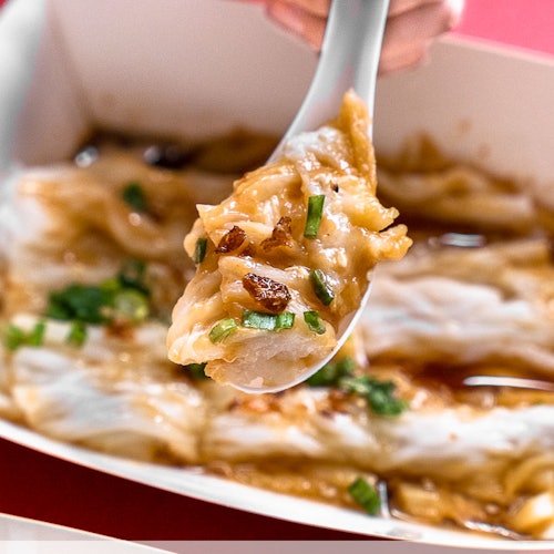 A close-up of a spoon holding a portion of rice noodles with sauce, garnished with green onions and bits of meat.