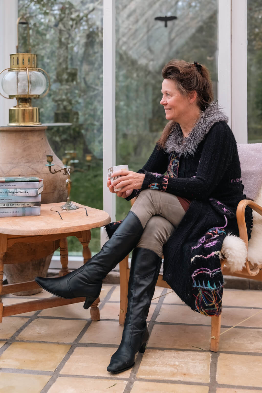 Billede af Suzi, som sidder i udestuen med en kop i hånden, mens hun kigger ud til siden. Ved siden er der en stak med hendes bøger og et antik lysestage på et lille bord.