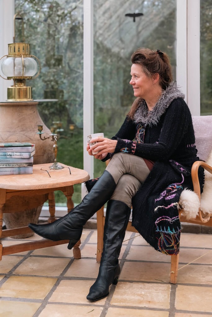 Billede af Suzi, som sidder i udestuen med en kop i hånden, mens hun kigger ud til siden. Ved siden er der en stak med hendes bøger og et antik lysestage på et lille bord.