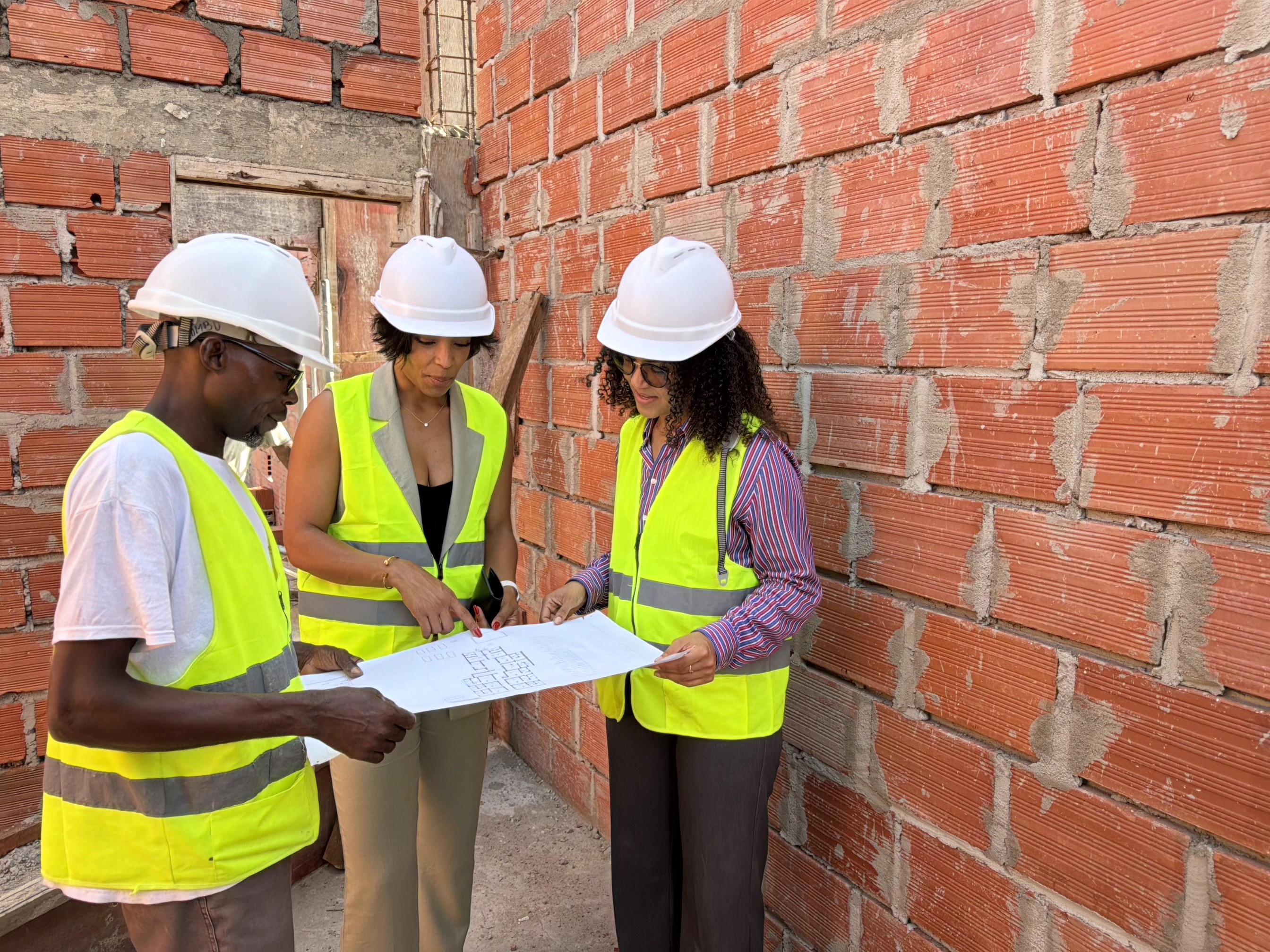 A building under construction is surrounded by scaffolding, with cranes in the background. A city skyline and waterfront are visible at sunset.