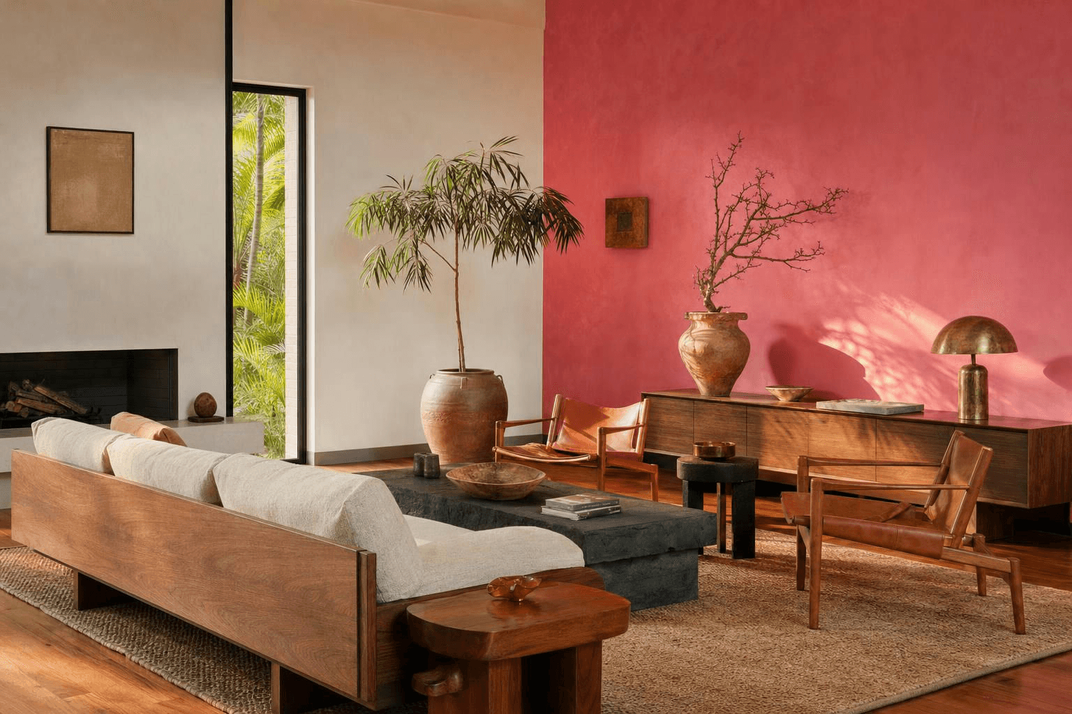 Living room with large pink accent wall, wood furniture, cream sofa, potted plants, woven rug, and decorative pottery.
