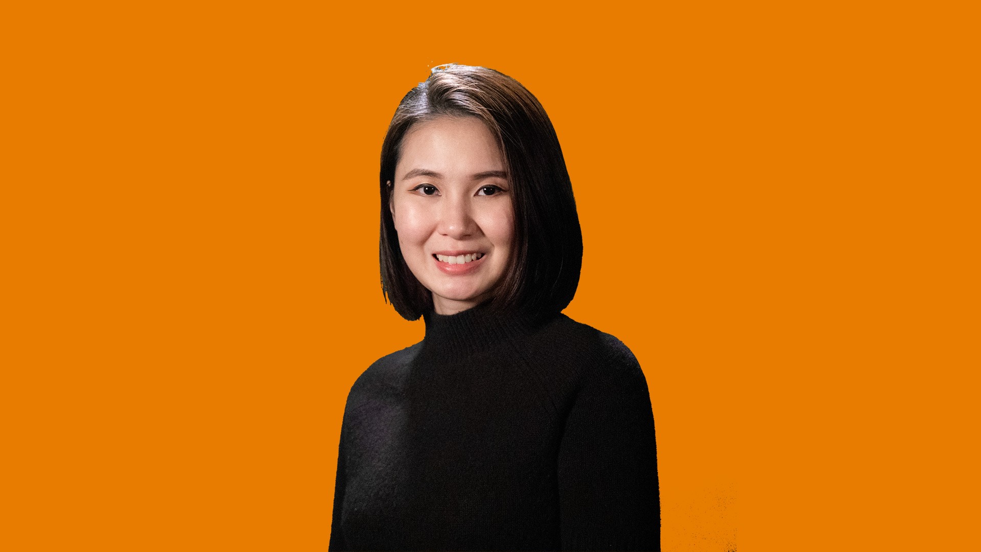 A woman with shoulder-length dark brown hair is centered against an orange background. She wears a black turtleneck sweater and smiles at the camera.