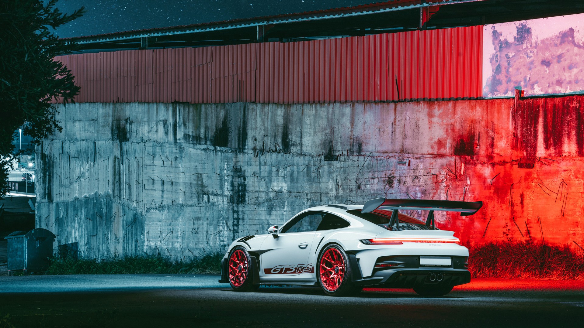 White Porsche 911 GT3 RS (992) with red wheels, shot from the rear three-quarter at night against a weathered concrete wall lit half in cool blue and half in deep red, swan-neck rear wing and centre-exit exhausts in frame, in an industrial setting in Nicosia.
