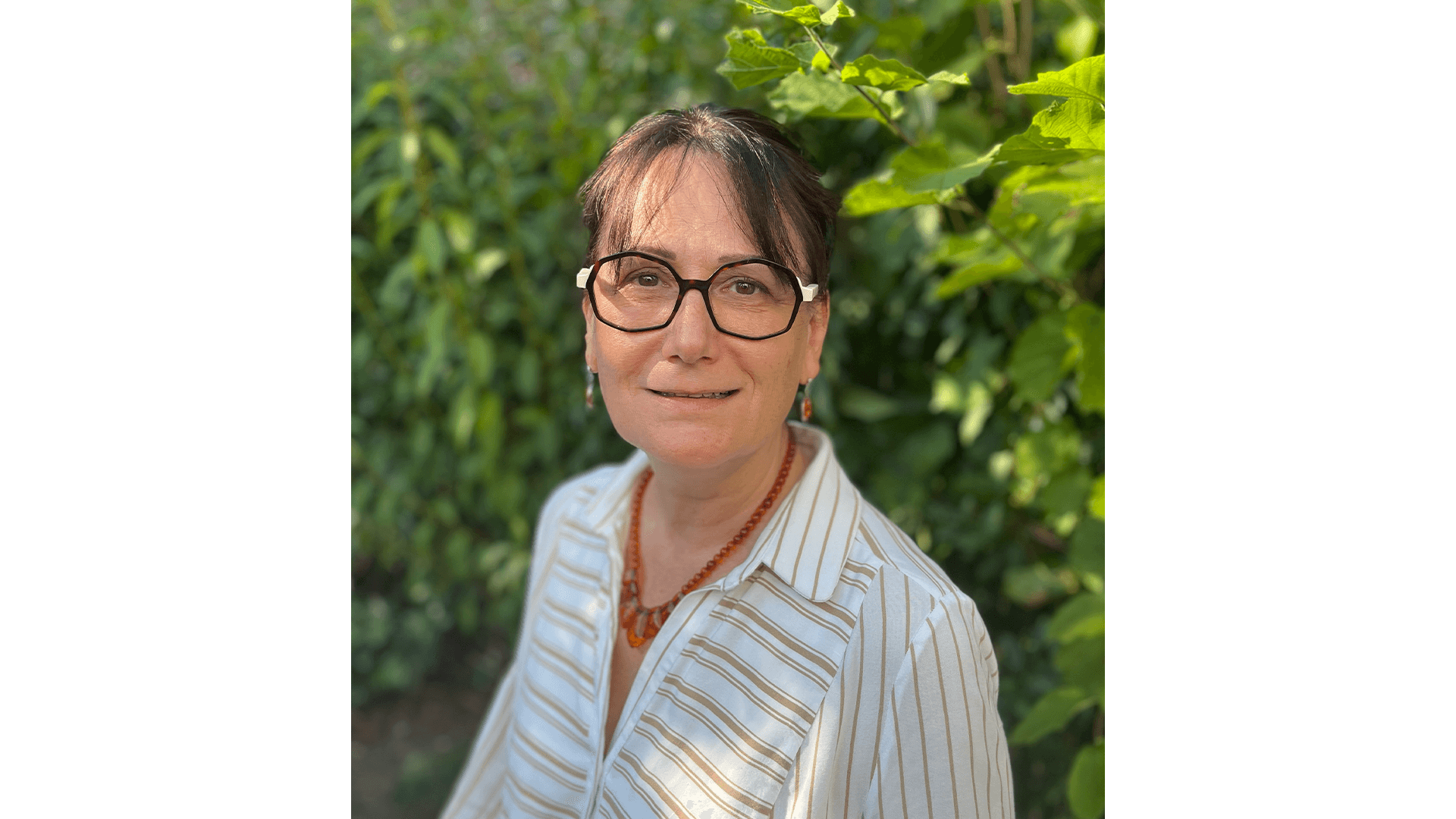 Smiling female therapist with short gray hair, wearing a brown top and a purple shawl, surrounded by a cozy background.
