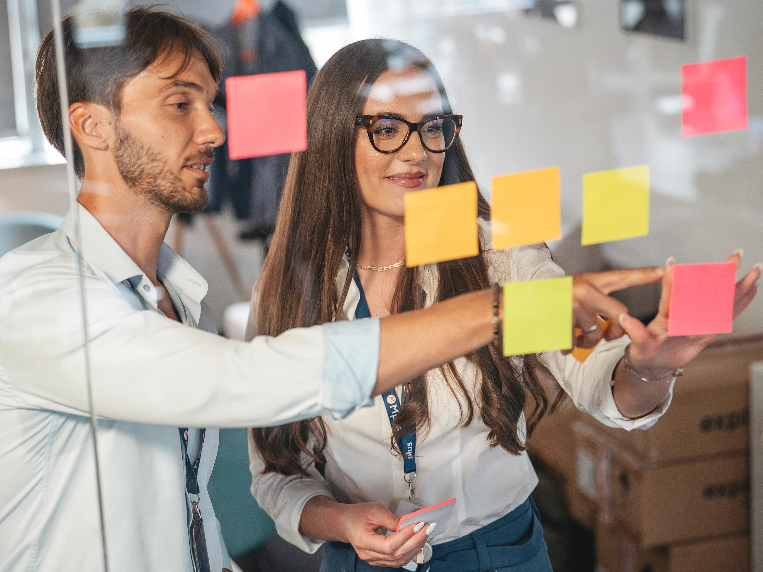 Two colleagues brainstorming and placing sticky notes on a glass board.