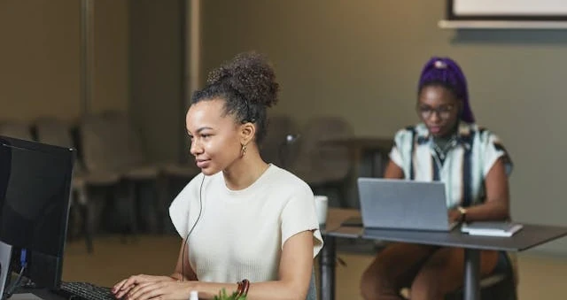 Mulher trabalhando num computador de mesa num escritório, com uma colega ao laptop ao fundo