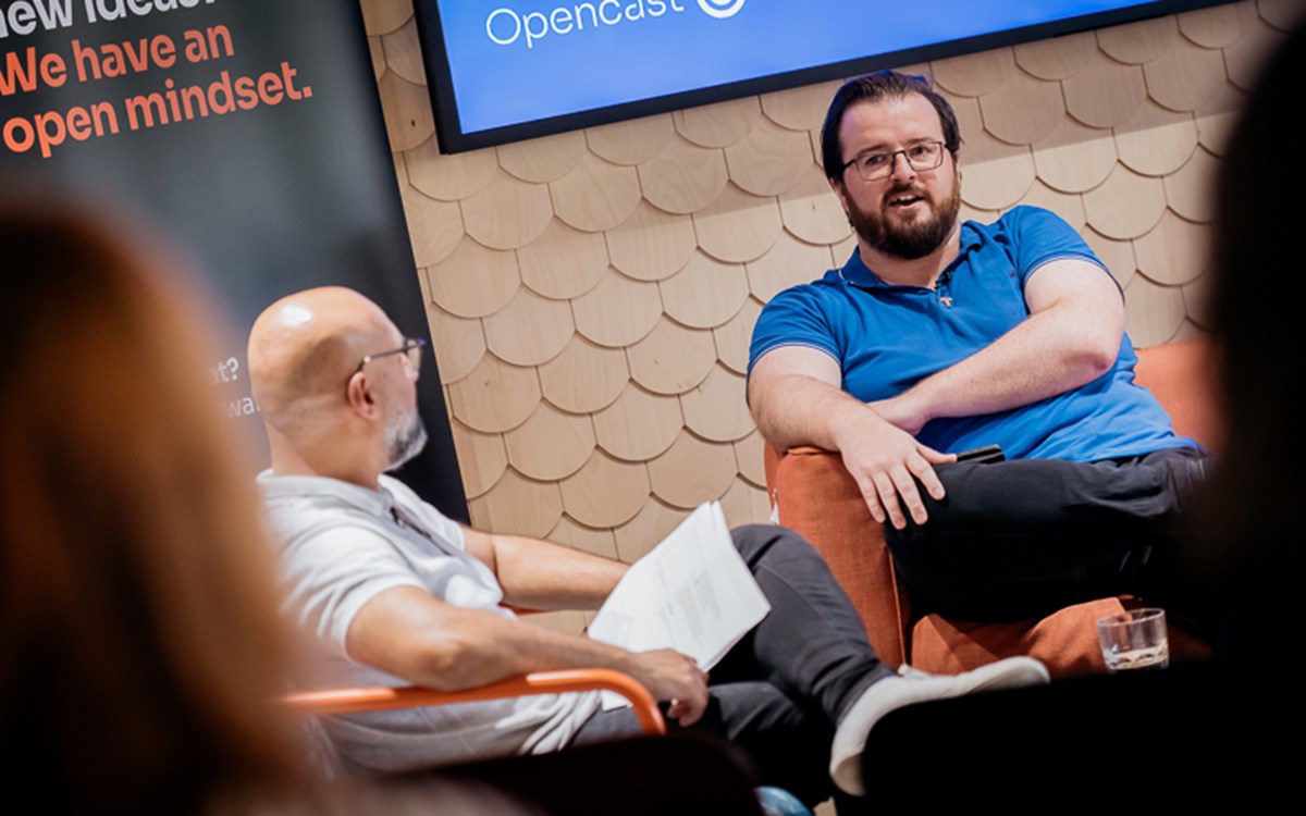 Two people seated indoors talking, with Opencast logo on screen and a banner about open mindset in the background.