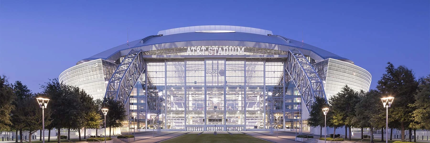 Exterior view of AT&T Stadium at dusk with illuminated glass facade and large steel arches framing the entrance.
