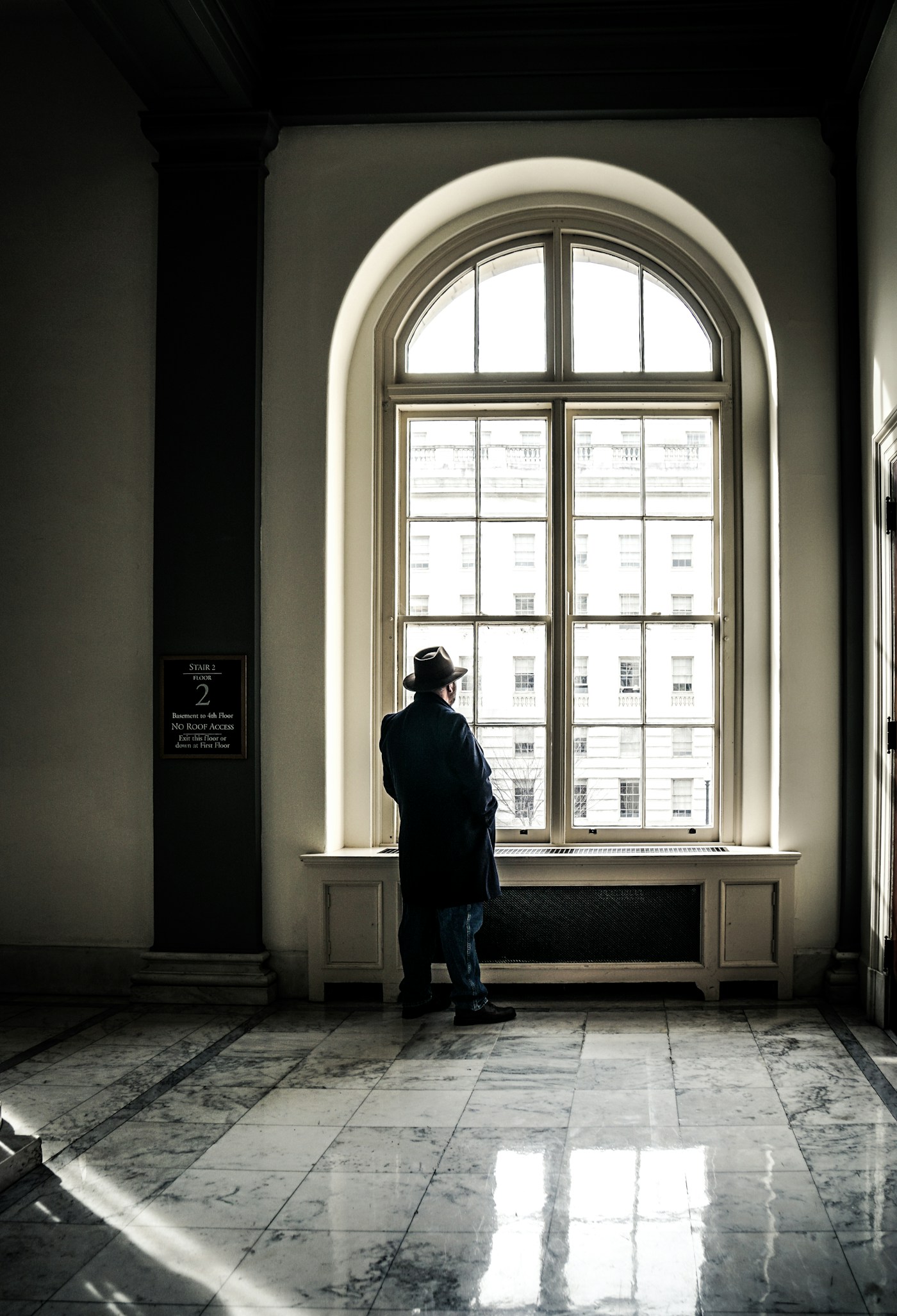 A lone figure standing at a tall arched window in a marble-floored hall