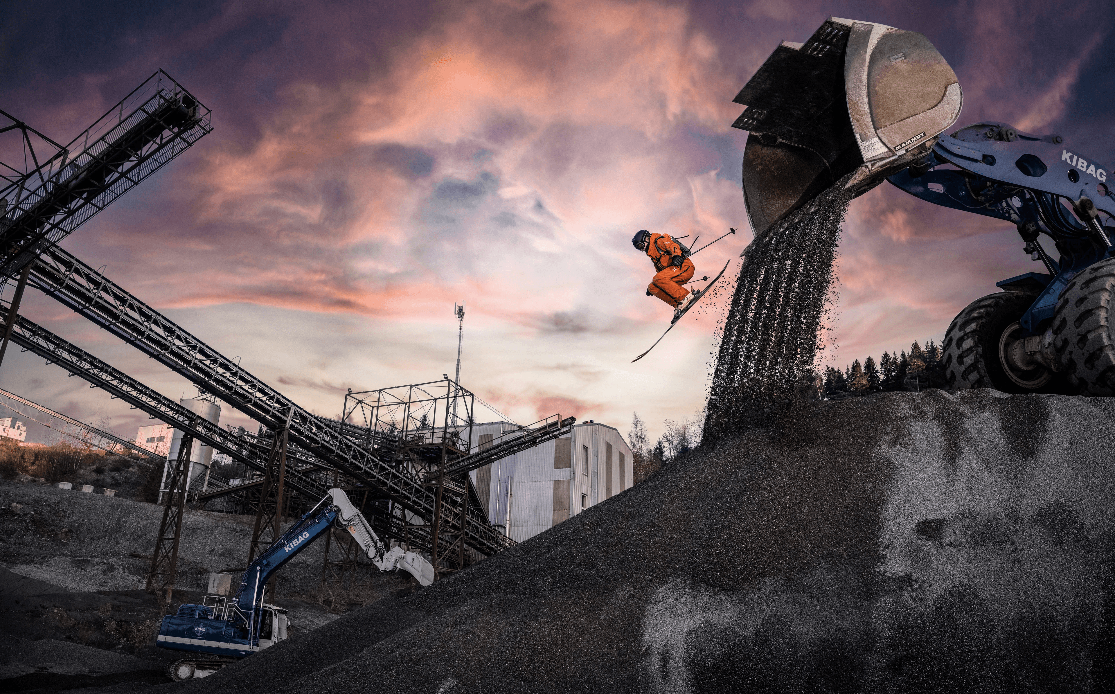 A skier in an orange outfit jumps off a gravel mound beneath an excavator at an industrial quarry during sunset. The image combines action sports and industrial machinery in a dramatic commercial photography scene created for Mammut by Rainer Eder.