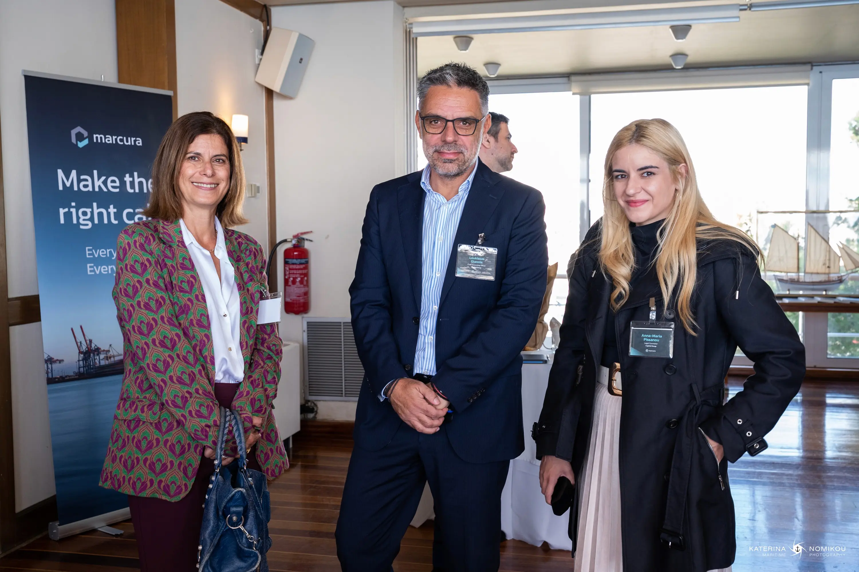 Three professionals standing together indoors, smiling, with a window and a promotional banner in the background.