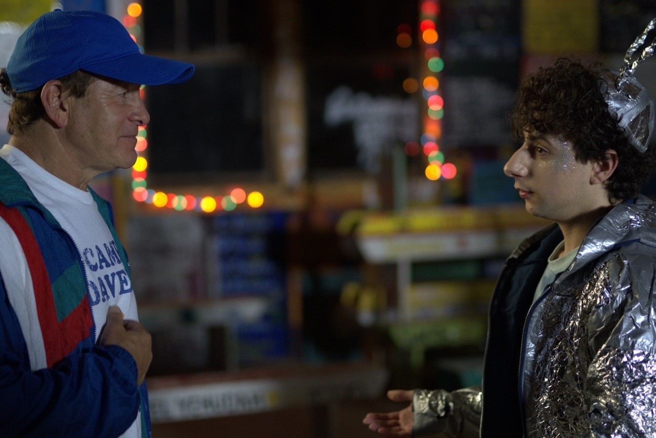 Still from The Floaters: A man in a baseball cap and a colorful jacket talks to a younger person dressed in a shiny, foil-covered costume with a handmade antenna.