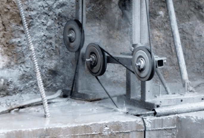 A diamond wire saw machine in operation, with the wire passing through two pulleys while cutting through a stone block. The machine is creating fine dust, indicating the precision of the cutting process.